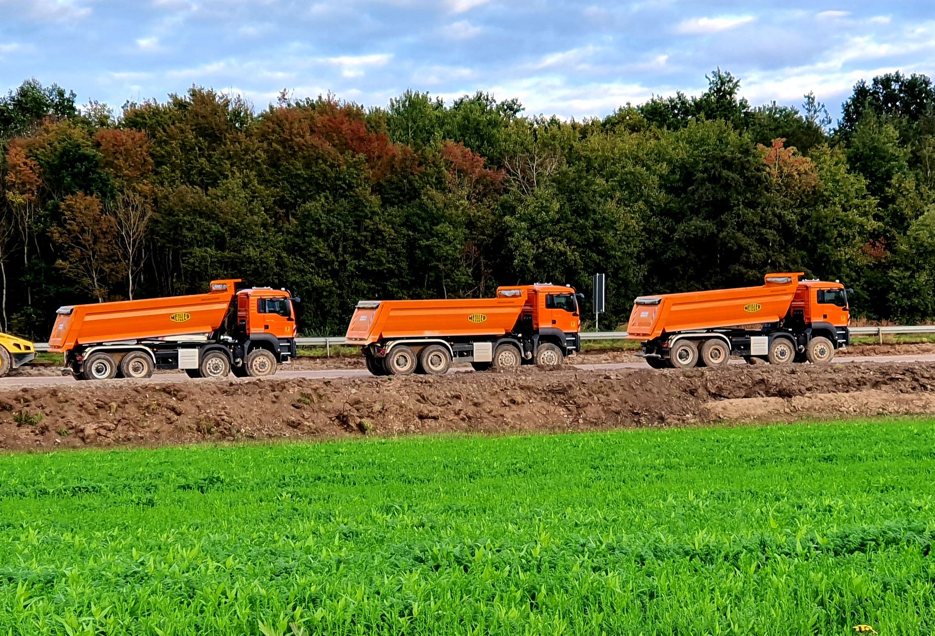 Three orange dump trucks parked at an outdoor construction site beside green grass fields. — Variables To Consider Before Buy